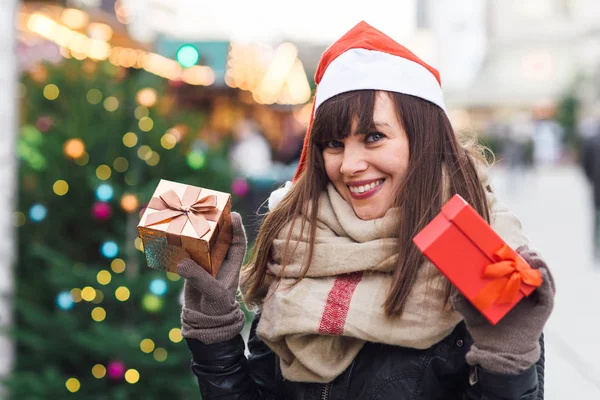 Beautiful woman in santa hat holding  two present boxes on christmas market — Stock Photo, Image