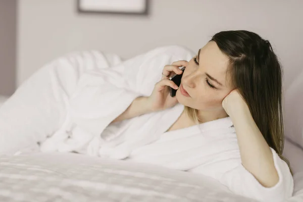 Hermosa mujer acostada en la cama y hablando en el teléfono inteligente en la habitación de hotel —  Fotos de Stock