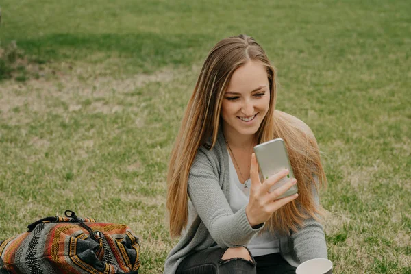 Ung flicka sitter på gräset med smart telefon och gör en selfy — Stockfoto