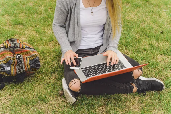 Jovem sentada na grama com telefone inteligente e laptop — Fotografia de Stock