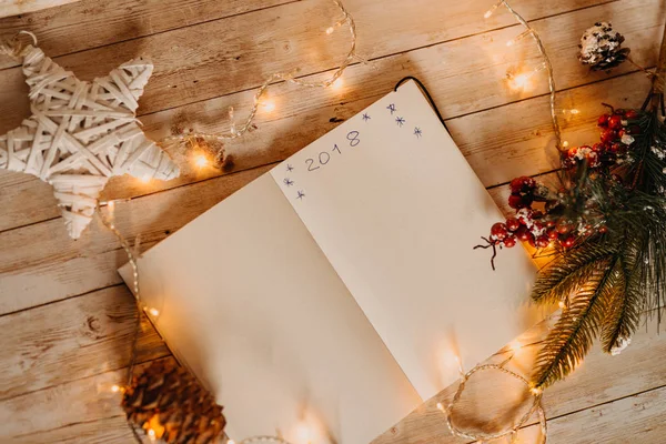 2018 written on opened notebook. top view of Christmas and new year decorations on wooden desk with lights — Stock Photo, Image