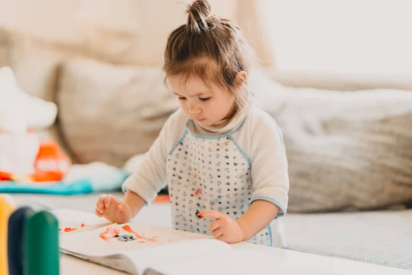 Toddler dziewczynka w rysunku fartuch zwraca palce w domu — Zdjęcie stockowe