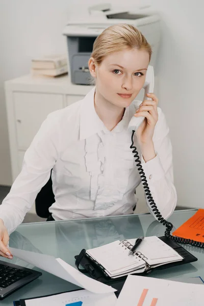 Vacker kvinna i office fungerar vid skrivbord, skriva i planner och talar på fast telefon — Stockfoto