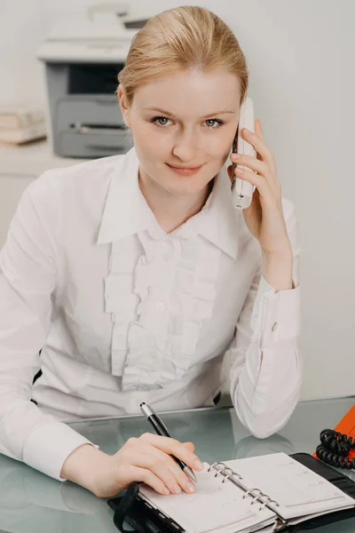 Schöne Frau im Büro arbeitet am Schreibtisch, schreibt im Planer und spricht am Festnetztelefon — Stockfoto