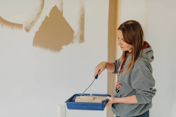 young woman painting wall with a roller and holding paint tray