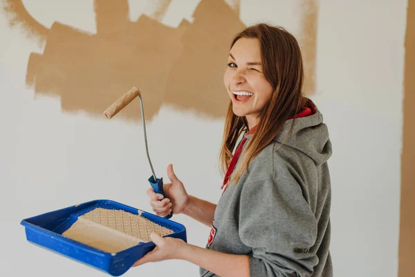 young woman painting wall with a roller and holding paint tray