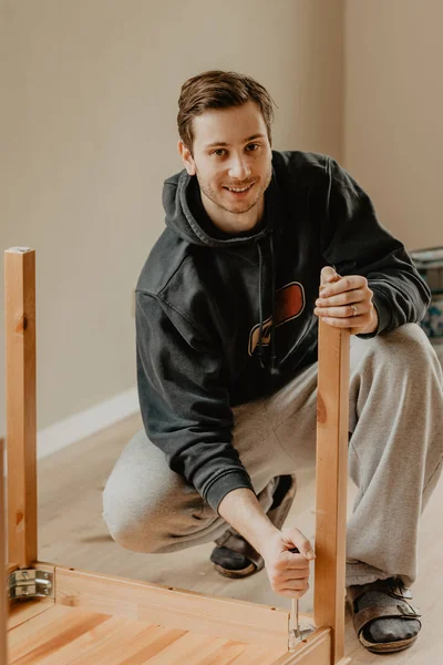 Hombre atornilla las piernas a una mesa de madera mirando en cámara y sonrisas —  Fotos de Stock