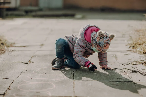チョークでアスファルトに描かれた顔のマスクの幼児の女の子は 屋外で寒い天候だけで遊んで — ストック写真