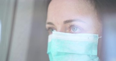 Tıbbi koruyucu maskeli yakın plan kadın pencereden dışarı bakıyor. Coronavirus Karantina Konsepti. Dışarıdaki kadın. Karantinaya alın. Koruyucu maske solunum cihazı