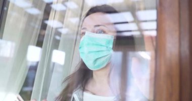 Tıbbi koruyucu maskeli bir kadın pencereden dışarı bakıyor. Coronavirus Karantina Konsepti. Dışarıdaki kadın. Karantinaya alın. Koruyucu maske solunum cihazı