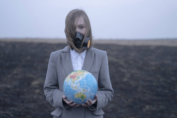 A student girl in a protective mask or a respirator holds a globe in her hands. on the background of earth in smoke and fire. burning grass leaves emissions Earth conservation concept, disaster