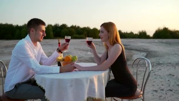 若い夫婦が自然とテーブルに座っています サンセットの砂浜でロマンチックなデートを ワインを飲むのが大好き笑顔話している 屋外だ 黒のドレスを着た赤い髪の少女 若い男 — ストック動画