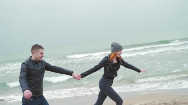 冬のビーチを一緒に走る楽しいカップルを愛しています 男と女の赤毛 黒い服を着て 冷たい雨笑顔の手を握って飛び跳ねる 海や海の岸です スローモーション — ストック動画