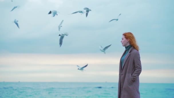 一个红头发的女孩 站在码头上 冬天或秋天 寒冷的天气 鸟儿和海鸥在飞翔 穿着外套 慢动作 寂寞的女人 — 图库视频影像