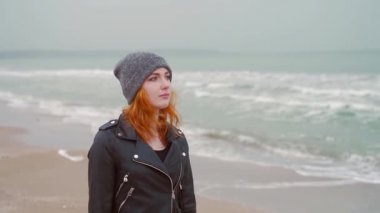 Genç kızıl saçlının portresi. Siyah elbiseli kızıl saçlı kız deniz kıyısında yürüyor, okyanus kıyısında soğuk hava, yavaş çekim, kış ya da sonbahar, büyük dalgalar, rüzgar, koyu deri