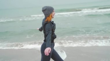 Siyah elbiseli ve deri ceketli genç kız. Kaçar, okyanus ya da deniz kenarındaki kameraya güler. Kış ya da sonbahar, soğuk hava, kum dalgalarının arka planına karşı ağır çekim. Mutlu.