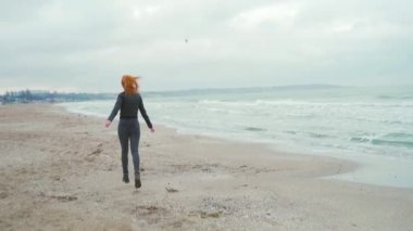 Siyah elbiseli ve deri ceketli genç kız. Kaçar, okyanus ya da deniz kenarındaki kameraya güler. Kış ya da sonbahar, soğuk hava, kum dalgalarının arka planına karşı ağır çekim. Mutlu.