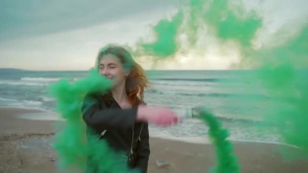 年轻的红头发女孩 手里拿着烟雾弹在海边奔跑 绿色的烟雾 黑色的衣服 在海洋的背景上 冬天或秋天 寒冷的天气缓慢的运动对着摄像机笑 — 图库视频影像