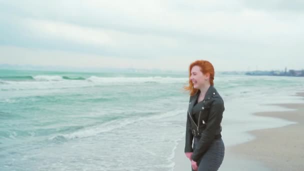 黒の服と革のジャケットの少女 渦は海や海のそばのカメラで笑います 冬か秋か寒気かゆっくりとした動き砂の波を背景に — ストック動画