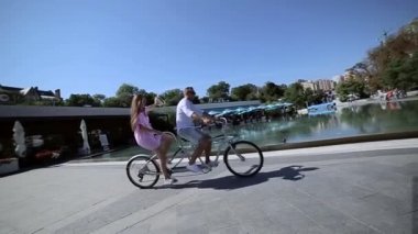 Genç, eğlenceli çift bisikleti. Genç, neşeli bir adam ve çift motorlu bir kız. Havuz kenarındaki yaza aşığım. Güneşli hava, pembe elbise beyaz gömlek, kot şort ve şapka. Gülümsüyor