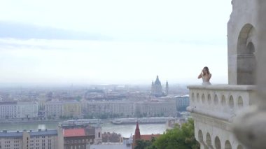 Gelin, arka plandaki eski kalenin balkonunda duruyor. Eski şehir ve nehir manzarası, yavaş çekim, eski mimarinin terasında genç güzel bir kız, Budapeşte, Bastion, Avrupa uzay metninin kopyası.