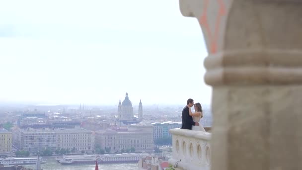 花嫁は古い城のバルコニーやテラスに立って抱き合ってキスをします 旧市街の風景を背景に 川の眺め スローモーション ブダペスト 仏ヨーロッパ コピースペーステキスト — ストック動画