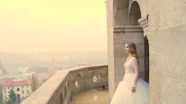 美丽的仙女公主在古城和河流的背景下俯瞰着一座精美古城堡的阳台或阳台 穿着白色婚纱的新娘 慢动作 — 图库视频影像