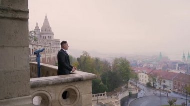 Siyah takım elbiseli, ceketli genç çekici bir adamın portresi eski şehir arka planında, şatoda, taş bina mimarisinde kemer ve sütunlarla gider. Lüks, zarif, sofistike, erkek, erkek. 