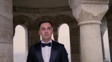Siyah takım elbiseli, ceketli genç çekici bir adamın portresi eski şehir arka planında, şatoda, taş bina mimarisinde kemer ve sütunlarla gider. Lüks, zarif, sofistike, erkek, erkek. 