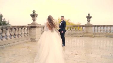 Gelin damada koşar, eski kalenin meydanında, eski kasabanın beyaz arka planı, uzun açık renk saçlar. Bali Malikanesi ve Baluster Sarayı. Yavaş çekim.