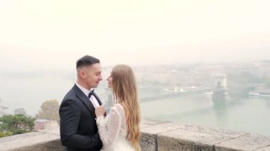 Eski şehir manzarasının arka planında, nehrin, köprünün, mimarinin arka planında sarılan bir çift. Avrupa. Sarışın, beyaz elbiseli ve damadı siyah olan çekici bir gelin.