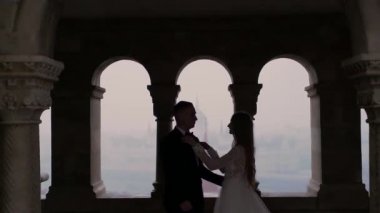 Eski şehir manzarasının arka planında, nehrin, köprünün, mimarinin arka planında sarılan bir çift. Avrupa. Sarışın, beyaz elbiseli ve damadı siyah olan çekici bir gelin.