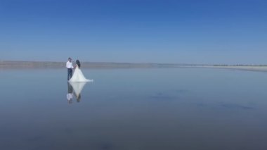 Gelin ve damat suya yansıdı. Genç bir düğün çifti, yazın denizin ortasında beyaz bir elbiseyle, gözlüklü, ayna görüntüsüyle dikiliyor. El ele tutuşun Hava görünümü