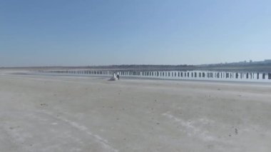 Gelin ve damat göl ya da nehir kıyısında beyaz gelinlik içinde kol kola yürüyorlar. Beyaz kum ve mavi suyla. Arka planda deniz ya da okyanus. Hava görünümü