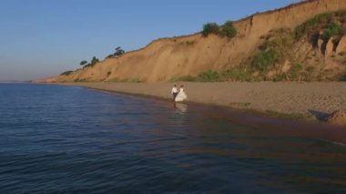Genç gelin ve damat, gün doğumunda deniz kenarında beyaz bir gelinlikle el ele yürüyorlar. Gün batımı. Mavi su, martı dalgaları, yaz. Aşıklar elden gider. Hava görüntüsü. Kayaların arkaplanı 