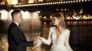 Genç bir çift, beyaz elbiseli gelin ve damat, dans ediyorlar, el ele tutuşuyorlar, sarılıyorlar, eski şehrin, nehrin ve büyük köprünün arka planında. Bokeh ışıkları. Işık. İyi akşamlar. Yavaş çekim.