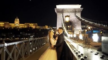 Portre. Düğün çifti, gelin ve damat beyaz elbiseli, sarılıyorlar, eski şehrin, nehrin ve büyük köprünün arka planında. Bokeh ışıkları. Işık. İyi akşamlar. Ağır çekim. moda, lüks aşıklar