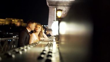 Portre. Düğün çifti, gelin ve damat beyaz elbiseli, sarılıyorlar, eski şehrin, nehrin ve büyük köprünün arka planında. Bokeh ışıkları. Işık. İyi akşamlar. Ağır çekim. moda, lüks aşıklar