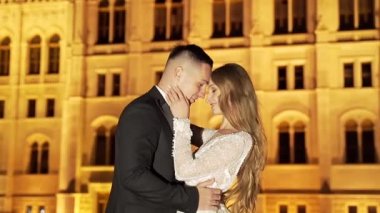 Portre. Düğün çifti, gelin ve damat beyaz elbiseli, sarılıyorlar, eski şehrin, nehrin ve büyük köprünün arka planında. Bokeh ışıkları. Işık. İyi akşamlar. Ağır çekim. moda, lüks aşıklar