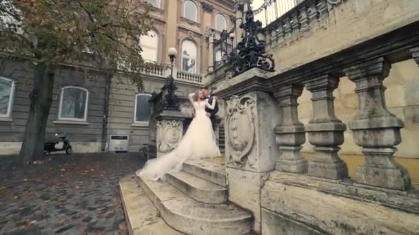 年轻的新婚夫妇依偎在旧城广场的楼梯上 在一座古老的石头建筑的背景下 新娘拥抱新郎 爱的故事 慢动作 — 图库视频影像