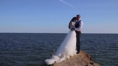 Genç bir düğün çifti tepenin kenarında, deniz ya da okyanusa karşı duruyor, gelin ve damat el ele tutuşuyor, mavi gökyüzü uçan kuş martıları, kum, kaya, beyaz elbise, siluet