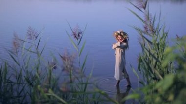 Kafasında çelenk olan keten renginde elbisesi olan genç sarışın kız suda duruyor ve dans ediyor. Klasik ulusal kostüm, Slav. Ivan Kupala. Etnik milli halk. Bir kadın yürür