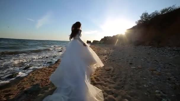 白いウェディングドレスを着た長髪の魅力的な女の子だけが海岸沿いを歩いています 花嫁は日没時に走ります 青い海を背景に 女性のスローモーション セクシーな女性 豪華な 上の幸せな笑顔 — ストック動画