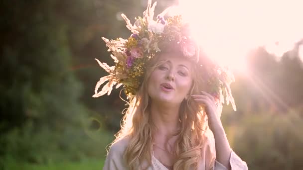 頭の上に花や植物の花輪を持つ若いウクライナの少女の肖像画 金髪の長髪の金髪の女性が日光の背景で歌います 自然は夏です 笑顔だ 閉じろ — ストック動画