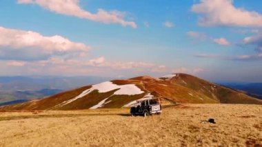 SUV bir dağın tepesinde duruyor ya da sürüyor. Dağların sırtı boyunca zorlu bir yolculuk. Mavi ve altın dağ manzaraları, kardaki tepeler. Vahşi doğa arazi aracı, araba..