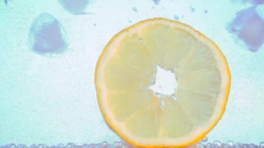 Kapatın. Bir dilim limon ya da portakal, buzlu karbonatlı soda suyuna düşer. Bir bardak maden suyunda. Ağır çekim. Makro. açık mavi beyaz arkaplan.