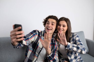 Kanepede selfie çekerken gülümseyen genç çift el sallıyor.