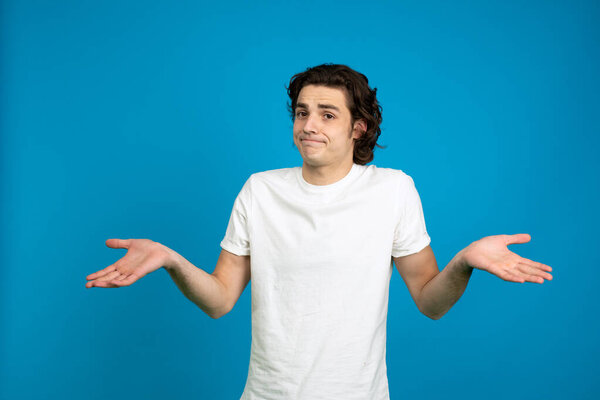 Front view of young man in white t-shirt shrugging isolated on blue background