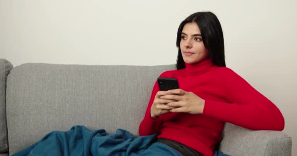 Bruneta relaxuje na gauči pomocí chytrého telefonu v domě. Holka píše sms, seznam případů. Šťastný pohled do kamery. Indoors — Stock video