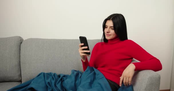 Telefondaki videoyu izleyen mutlu kız. Sohbet etmek ve duygularını göstermek. Esmer, evdeki oturma odasında kanepede oturuyor. Sosyal ağdaki sayfaya bak. — Stok video